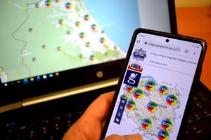 Na zdjęciu trzymany w ręku telefon, na ekranie którego widoczna jest mapa Polski otworzona w aplikacji Krajowa Mapa Zagrożeń Bezpieczeństwa. W tle laptop i na jego ekranie mapa Polski otwarta w tej samej aplikacji