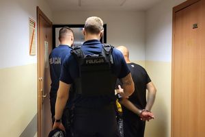 Umundurowani policjanci wprowadzają do pomieszczenia mężczyznę, który ma założone kajdanki na ręce trzymane z tyłu.