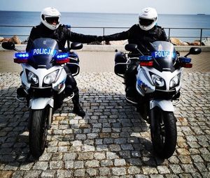 Dwaj umundurowani policjanci siedzą na motocyklach i przybijają sobie piąstkę. Za nimi w tle woda