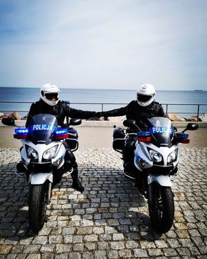 Dwaj umundurowani policjanci siedzą na motocyklach i przybijają sobie piąstkę. Za nimi w tle woda