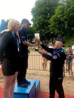 Umundurowany policjant wręcza umundurowanej poicjantce puchar. Policjantka razem z inną kobietą stoją na podium dla zawodników.  W tle dzieci i dorośli obserwujący dekorację zawodniczek.