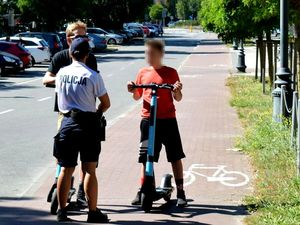 Umundurowana policjantka rozmawia z mężczyzną i chłopcem, którzy trzymają hulajnogi elektryczne. Za nimi parking samochodowy, na którym zaparkowane są pojazdy. Zdjęcie archiwalne.