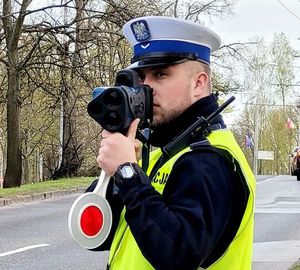 Umundurowany policjant trzyma w rękach urządzenie i mierzy prędkość pojazdów jadących ulicą