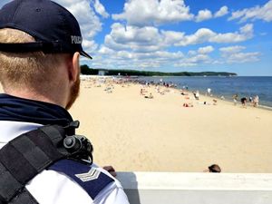 Na zdjęciu fragment sylwetki umundurowanego policjanta, a za nim w tle plaża nad wodą i wypoczywający na plaży ludzie