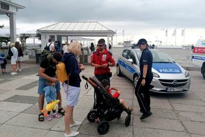 Na zdjęciu policjantka, ratownik i grupa osób. Za nimi radiowóz policyjny, a w tle inne osoby