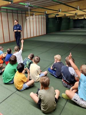 Na zdjęciu pomieszczenie strzelnicy. Na podłodze siedzą dzieci, a przed nimi stoi policjantka