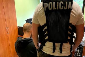 Na zdjęciu policjant w stroju cywilnym i z założoną kamizelką z napisem POLICJA, a obok niego siedzi na krześle mężczyzna w stroju cywilnym