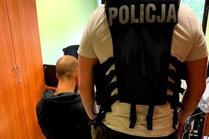 Na zdjęciu policjant w stroju cywilnym i z założoną kamizelką z napisem POLICJA. Obok niego na krezśle siedzi zatrzymany mężczyzna