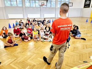 Na zdjęciu ratownik wodny, a za nim grupa dzieci siedzących na parkiecie hali