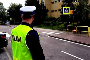 Na zdjęciu policjant, który stoi przy ulicy i patrzy na przejście dla pieszych. W tle budynek szkoły.