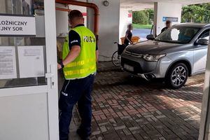 Na zdjęciu policjant, który trzyma otwarte drzwi na oddział szpitalny. Obok policjanta znajduje się samochód, w którym siedzi kobieta z zasłoniętą twarzą, a przy aucie stoi kobieta i mężczyzna z zasłoniętymi twarzami