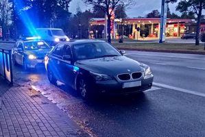 Pora nocna. Na zdjęciu ulica, a na niej stoi samochód bmw i za nim radiowóz policyjny. W tle widoczny kolejny samochód, który jedzie ulicą.