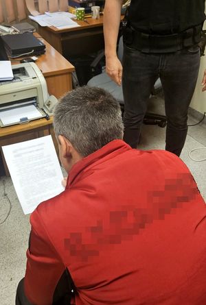 Na zdjęciu zatrzymany mężczyzna, który siedzi na krześle i czyta dokument. Za nim w tle stoi policjant w stroju cywilnym, u którego przy pasie widać broń w kaburze