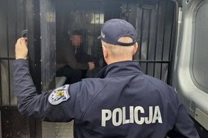 Na zdjęciu policjant, który zamyka pomieszczenie w radiowozie służące do przewozu osób zatrzymanych. W tle za policjantem w pomieszczeniu tym widoczny zatrzymany mężczyzna.