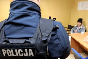 Na zdjęciu pomieszczenie służbowe. Na pierwszym planie widać plecy policjanta, który ma załóżoną kamizelkę z napisem POLICJA, a za nim w tle na krześle siedzi kobieta.