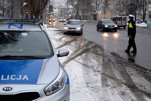 Na zdjęciu radiowóz i policjant, który zatrzymuje do kontroli kierującego samochodem. Obok po ulicy jadą inne samochody, a na drzewach i poboczu leży śnieg.