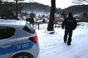 Na zdjęciu radiowóz i policjant, który idzie w stronę ogródków działkowych. Sceneria zimowa, na ziemi leży śnieg.