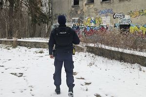 Umundurowany policjant idzie w stronę opuszczonego budynku. Na ziemi leży śnieg