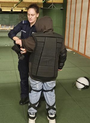 Na zdjęciu policjantka oraz uczennica. Uczennica ma założoną  na sobie kamizelkę policyjną i ochraniacze na nogach, a policjantka zakłada jej ochraniacz na rękę