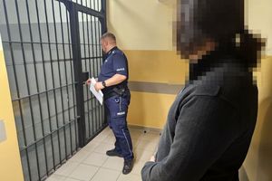 Na zdjęciu po prawej stronie znajduje się zatrzymany mężczyzna. Za nim w tle widoczny umundurowany policjant, który otwiera okratowane drzwi do pomieszczenia dla osób zatrzymanych