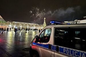 Na zdjęciu radiowóz. Za nim w tle osoby spacerujące po placu, choinka świąteczna i fajerwerki na niebie