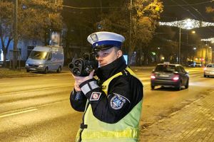 Pora nocna. Na zdjęciu policjant ruchu drogowego, który stoi przy ulicy za pomocą urządzenia trzymanego w rękach mierzy prędkość pojazdów jadących ulicą. W tle pojazdy jadące ulicą i jeden stojący na poboczu.