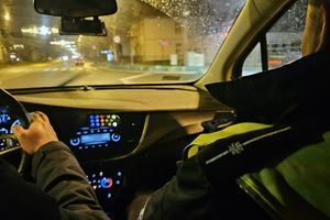 Na zdjęciu wnętrze radiowozu, a w nim siedzi dwóch policjantów i jadą ulicą. Na zewnątrz pora nocna