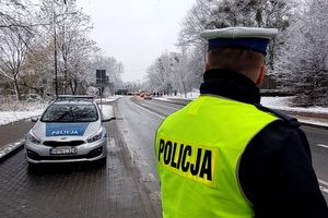 Pora zimowa, na drzewach i ziemi leży śnieg. Na zdjęciu policjant, który stoi przy ulicy. Obok niego stoi radiowóz, a w tle po ulicy jadą samochody