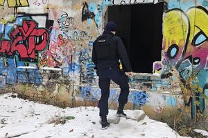 Umundurowany policjant idzie w stronę otworu w pustostanie. Na ścianie budynku znajdują się graffiti a na ziemi przy budynku leży śnieg