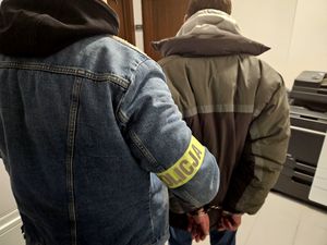 Na zdjęciu policjant w stroju cywilnym z założoną opaską na ramieniu z napisem POLICJA, który prowadzi zatrzymanego mężczyznę z założonymi kajdankami na rękach