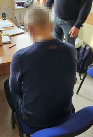 Na zdjęciu zatrzymany mężczyzna, który siedzi przy biurku. Za nim stoi policjant w stroju cywilnym