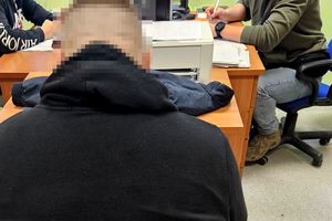Na zdjęciu zatrzymany mężczyzna, który siedzi na krześle w pomieszczeniu służbowym. Za nim przy biurkach siedzą policjanci w strojach cywilnych, którzy wykonują czynności z zatrzymanym