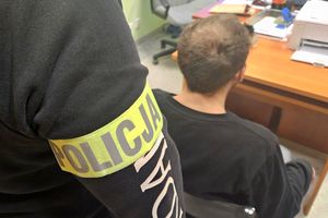 Na zdjęciu policjant w stroju cywilnym z założoną na ramieniu opaską z napisem POLICJA, a obok niego siedzi na krzśle zatrzymany mężczyzna