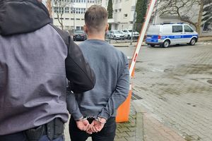 Na zdjęciu zatrzymany mężczyzna z założonymi na rękach kajdankami, którego prowadzi policjant w stroju cywilnym. W tle radiowóz policyjny i samochody cywilne