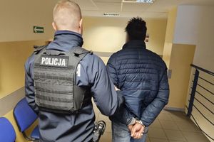 Policjant prowadzi zatrzymanego mężczyznę, który ma założone kajdanki na ręce trzymane z tyłu.