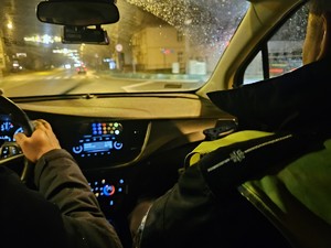 Na zdjęciu widać policjanta siedzącego na fotelu pasażera i częściowo widać policjanta kierującego autem. Jadą oni radiowozem. Zdjęcie zrobione w porze nocnej