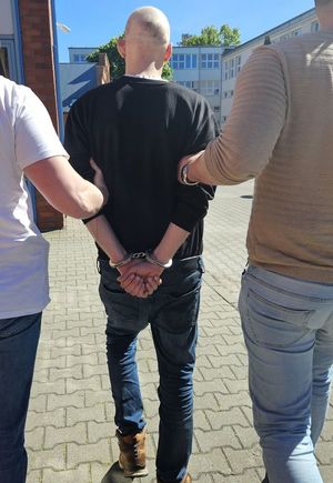 Na zdjęciu zatrzymany mężczyzna, który ma założone kajdanki na ręce trzymane z tyłu. Prowadzi go dwóch policjantów w strojach cywilnych.