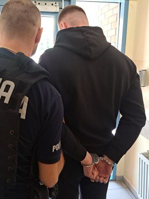 Na zdjęciu policjant i zatrzymany mężczyzna, który ma kajdanki założone na ręce trzymane z tyłu