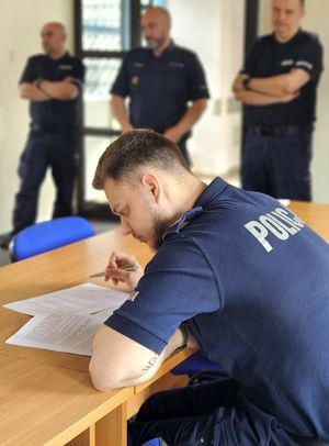 Na zdjęciu policjant siedzący przy biurku i rozwiązujący test. Za nim w tle trzech policjantów, którzy stoją