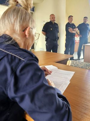 Na zdjęciu policjantka siedząca przy biurku i rozwiązująca test. Za nią w tle trzech policjantów, którzy stoją