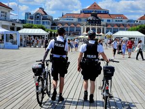 Na zdjęciu dwóch policjantów, którzy prowadzą rowery po Sopockim Molo. Za nimi w tle wiele osób i budynki