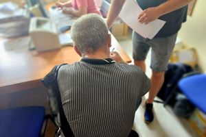 Na zdjęciu zatrzymany mężczyzna, który siedzi na krześle w pomieszczeniu służbowym. Obok mężczyzny stoi policjant w stroju cywilnym, a za nim w tle przy biurku siedzi policjant w stroju cywilnym.