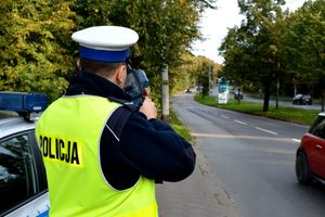 Na zdjęciu policjant, który trzyma w rękach urządzenie do pomiaru prędkość i i mierzy prędkość jadących ulicą samochodów