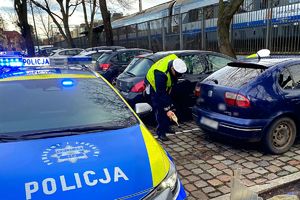 Na zdjęciu radiowóz, obok niego zaparkowane inne samochody i dwaj policjanci stojący przy jednym z tych samochodów. W tle pociąg