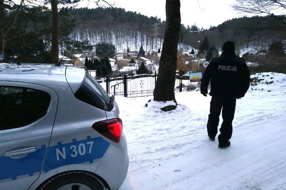 Na zdjęciu radiowóz, policjant i teren ogródków działkowych znajdujący się na skraju lasu. Aura zimowa, na ziemi leży śnieg